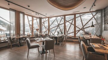 Ein modernes Restaurant mit großen Fenstern und Blick auf die Berge. Die Einrichtung ist stilvoll und gemütlich, mit Holz- und Stoffelementen.