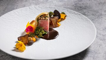Ein elegantes Gericht auf einem weißen Teller, bestehend aus Fleisch, Beilagen und dekorativen essbaren Blüten. Die Anordnung ist kunstvoll und ansprechend.