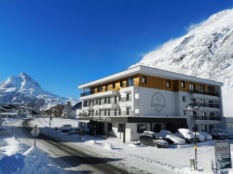 Ein modernes Hotel mit dem Namen „Zontaja“ in einer winterlichen Berglandschaft. Die Umgebung ist mit Schnee bedeckt und im Hintergrund sind majestätische Berge zu sehen.