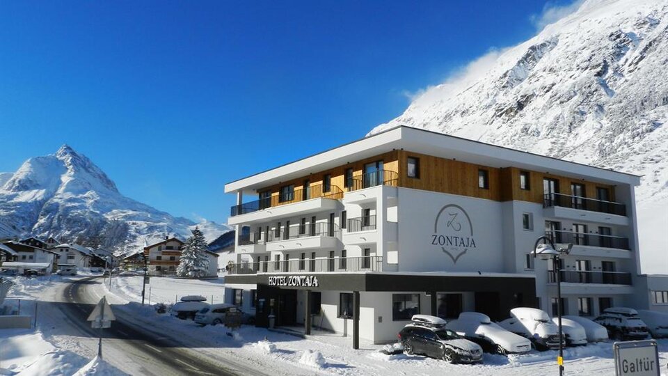 Ein modernes Hotel mit dem Namen „Zontaja“ in einer winterlichen Berglandschaft. Die Umgebung ist mit Schnee bedeckt und im Hintergrund sind majestätische Berge zu sehen.
