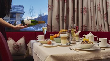 Ein gedeckter Tisch mit verschiedenen Frühstücksgerichten und einem Glas Sekt. Im Hintergrund sind Fenster mit Blick auf die Berge zu sehen.