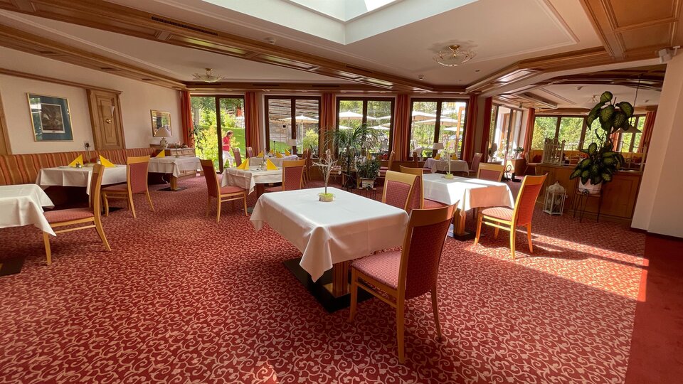Ein einladendes Restaurant mit eleganten Tischen und roten Teppichboden. Große Fenster sorgen für viel Tageslicht und eine angenehme Atmosphäre.
