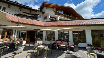 Eine gemütliche Außenterrasse eines Restaurants mit Sonnenschirmen und stilvollen Möbeln. Im Hintergrund ist ein modernes Gebäude mit Holzverzierungen und blühenden Fenstern.