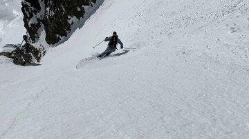 Ein Skifahrer fährt die steile, schneebedeckte Piste hinunter. Im Hintergrund sind Felsen und die verschneite Berglandschaft zu sehen.