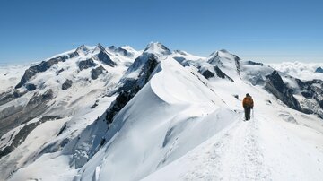 Ein Wanderer auf einem schneebedeckten Gipfel mit beeindruckenden Bergen im Hintergrund. Der Himmel ist klar und blau.
