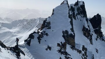 Eine beeindruckende Berglandschaft mit schneebedeckten Gipfeln. Im Vordergrund steht ein Wanderer auf einem schmalen Grat.