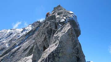 Ein steiler Felsen unter einem klaren blauen Himmel. Seil und Kletterausrüstung deuten auf Bergsteigen hin.
