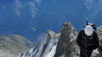 Ein Bergsteiger steht auf einem felsigen Gipfel mit schneebedeckten Bergen im Hintergrund. Der Himmel ist klar und blau, und die Landschaft ist majestätisch.
