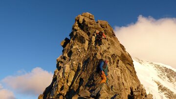 Zwei Kletterer steigen einen steilen Felsen bei Sonnenuntergang. Die Umgebung ist majestätisch mit schneebedeckten Bergen und wolkenfreiem Himmel.