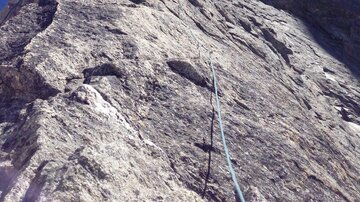 Eine steile Felswand unter einem klaren blauen Himmel. Das Sonnenlicht strahlt auf die Felsen, während Kletterer den Aufstieg beginnen.