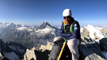 Ein Bergsteiger sitzt auf einem Gipfel und blickt auf eine beeindruckende Berglandschaft. Die Sonne scheint hell auf die verschneiten Gipfel im Hintergrund.