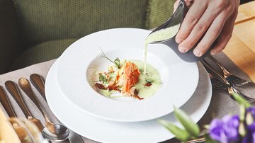 Ein elegantes Gericht wird auf einem weißen Teller serviert. Eine Hand gießt eine grüne Sauce über das Essen, das mit Kräutern garniert ist.