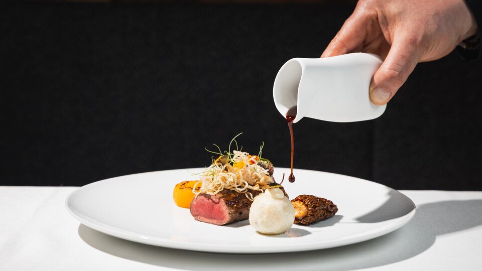 Eine stilvolle Speisepräsentation auf einem weißen Teller. Eine Hand gießt eine Sauce über das Gericht, das aus Fleisch, buntem Gemüse und Beilagen besteht.