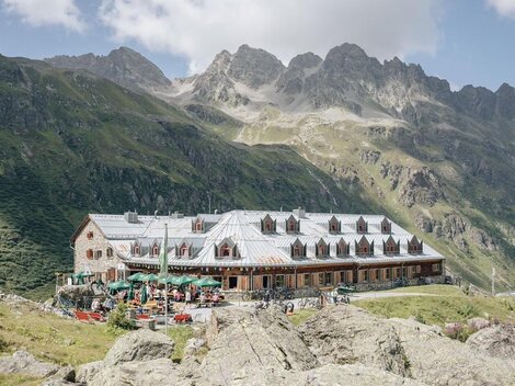 Eine gemütliche Berghütte mit Sitzgelegenheiten im Freien und vielen Gästen. Die Umgebung ist von beeindruckenden Bergen und einer klaren Bl sky umgeben.