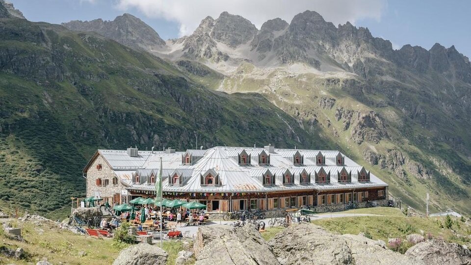 Eine gemütliche Berghütte mit Sitzgelegenheiten im Freien und vielen Gästen. Die Umgebung ist von beeindruckenden Bergen und einer klaren Bl sky umgeben.