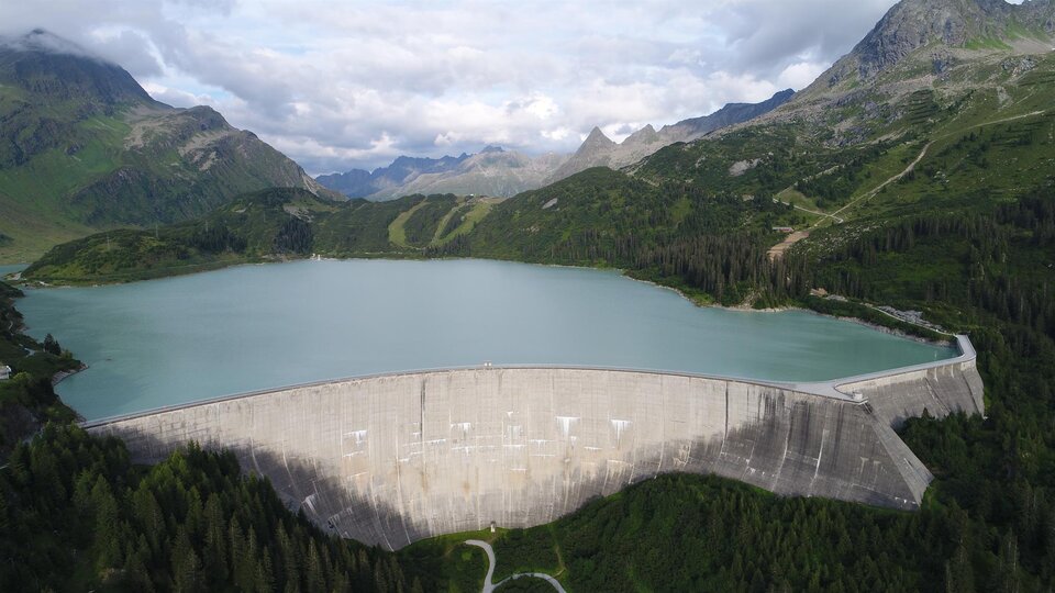 An impressive dam surrounded by green mountains and a clear lake. The scenery depicts a tranquil natural backdrop with a slightly cloudy sky.