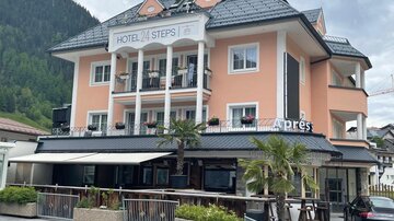 Ein modernes Hotel mit der Aufschrift "Hotel Steps" in einer malerischen Umgebung. Das Gebäude ist hell gestrichen und hat eine einladende Terrasse mit Pflanzen.