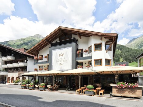 Ein gemütliches Hotel mit einem einladenden Außenbereich und Terrassen. Im Hintergrund sind grüne Berge und ein blauer Himmel zu sehen.