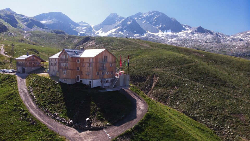 Ein modernes Gebäude auf einer grünen Anhöhe, umgeben von Bergen. Im Hintergrund sind schneebedeckte Gipfel und ein klarer blauer Himmel zu sehen.