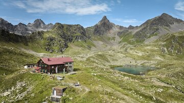 A picturesque mountain landscape with a mountain cabin and a clear lake. The surroundings are green and surrounded by impressive mountains.