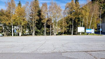 Eine leere Asphaltfläche mit herbstlichen Bäumen im Hintergrund. Neben den Bäumen stehen Informationsschilder.