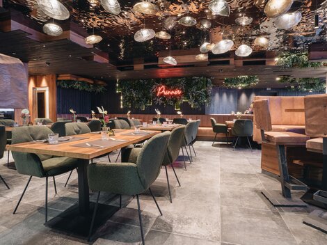 Ein modernes Restaurant mit gemütlichen Tischen und stilvollen Stühlen. Die Beleuchtung und Pflanzen schaffen eine einladende Atmosphäre.