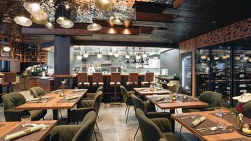 Ein elegantes Restaurant mit modernen Möbeln und warmem Licht. Die Tische sind schön gedeckt und eine offene Küche ist im Hintergrund sichtbar.