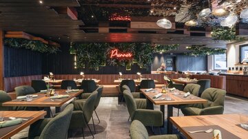 Ein modernes Restaurant mit eleganten Tischen und Stühlen. Der Raum ist mit Pflanzen und einer leuchtenden Neonschrift dekoriert.