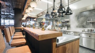 Ein modernes Restaurant mit einer offenen Küche und einer langen Holzbar. Die Einrichtung ist stilvoll und die Beleuchtung sorgt für eine einladende Atmosphäre.