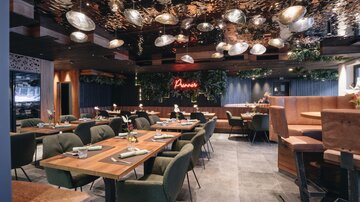 Ein modernes Restaurant mit stilvollen Holz- und Grüntönen. Die Tische sind elegant gedeckt und eine gemütliche Atmosphäre wird durch die Beleuchtung geschaffen.
