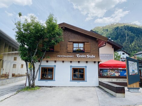 Ein gemütliches Restaurant namens "Tiroler Stube" mit Holzverkleidung und Fenstern. Im Hintergrund sind die Berge zu sehen.