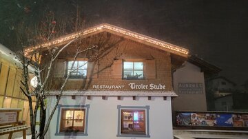 A cozy restaurant named "Tiroler Stube," illuminated by lights, stands in the snow. The scene conveys a wintry atmosphere at night.