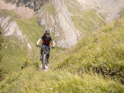 Ein Radfahrer fährt auf einem schmalen Weg durch die grüne Berglandschaft. Im Hintergrund sind felsige Berge und ein klarer Himmel zu sehen.