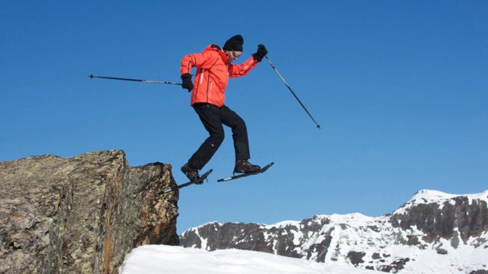 Ein Mann in leuchtend orangefarbener Jacke springt über einen Felsen im Schnee. Im Hintergrund sind schneebedeckte Berge und ein klarer blauer Himmel zu sehen.