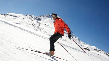 Eine Person in roter Ski-Ausrüstung fährt auf einer schneebedeckten Piste. Im Hintergrund sind schneebedeckte Berge und ein klarer blauer Himmel zu sehen.