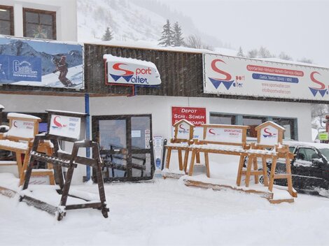 A sports store in winter with lots of snow in front of the door. The facade displays signs and wooden racks for ski equipment.