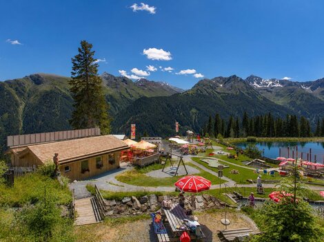 An alpine recreational facility set against an impressive mountain backdrop: In the foreground, a wooden terrace, sunbathing lawn, playground, and red sun umbrellas invite you to linger, while on the right, a clear mountain lake reflects the surrounding peaks. The bright wooden building, green meadows, and wide blue sky create a summery, family-friendly atmosphere in the heart of the Alps.