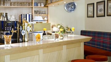Eine gemütliche Bar mit Holzregalen und einem Kaffeeautomaten. Auf der Theke stehen Getränke und dekorative Elemente, während die Sitzgelegenheiten rot gepolstert sind.