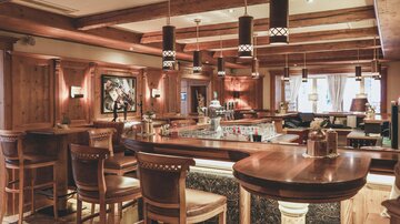 A cozy bar with wooden paneling and stylish stools. Various drink bottles are on the counter, and there is soft lighting.