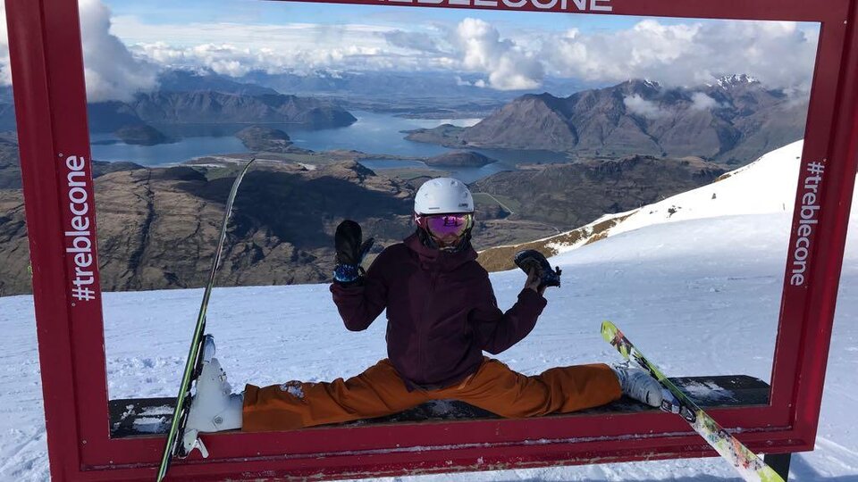 A skier is in a split position in a red frame. In the background, snow-covered mountains and a beautiful lake can be seen.