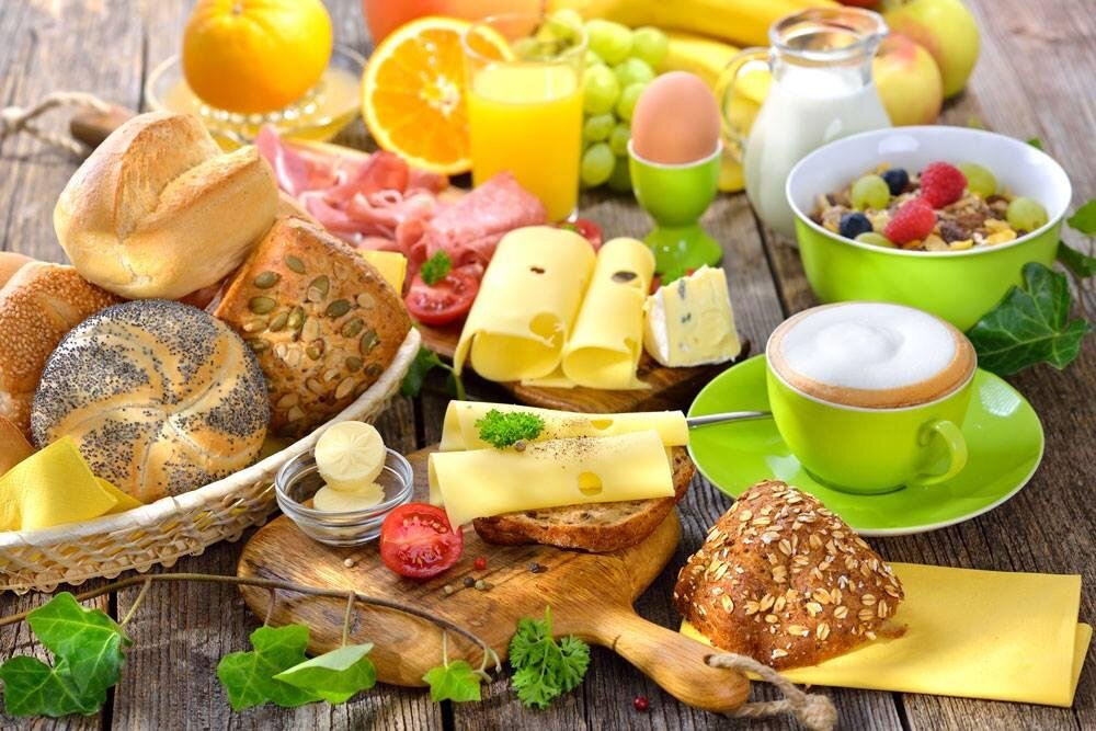 Ein reichhaltiges Frühstück mit verschiedenen Brotsorten, buntem Obst und Getränken. Dazu gibt es Käse und Joghurt in appetitlichen Schalen.