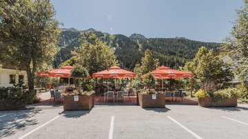 A cozy outdoor area with red umbrellas and chairs. In the background, the green mountains can be seen.