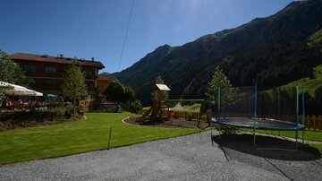 A beautiful garden with a playground and a trampoline. In the background, green mountains and a blue sky can be seen.