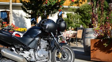 A black motorcycle is standing in a cozy outdoor area with flowers and tables. In the background, guests can be seen relaxing.