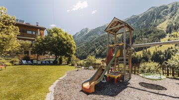 A playground with a slide is in the foreground. In the background, beautiful mountains and a green meadow can be seen.