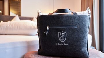 An elegant black bag lies on a stool in a modern room. In the background, a bed and curtains are visible.