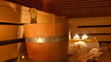 Ein Holzkorb steht in einer Sauna, umgeben von Holzbank und Kerzenlicht. Entspannte Atmosphäre mit Orchideenblüten für das Wohlfühl-Erlebnis.