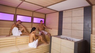 A modern sauna with a wooden bench and soft, colored lighting. Three people are relaxing in towel-like clothing.