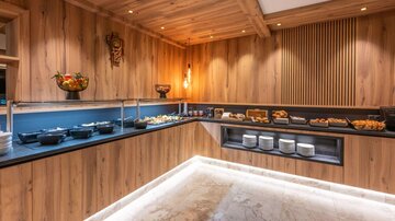 A stylish breakfast area with wood-paneled walls and elegant lighting. The counter offers a selection of various foods and drinks.
