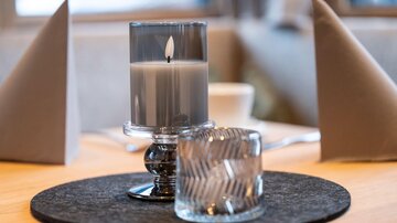 An elegant table with a candle in a glass and a decorative cup. In the background, gray napkins and a cozy atmosphere can be seen.
